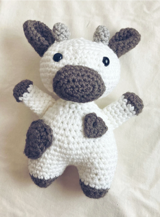 Handmade crochet cow plush toy in white and grey yarn with stitched details, sitting upright. Perfect for nurseries, plushie collections, or farm-themed gifts.